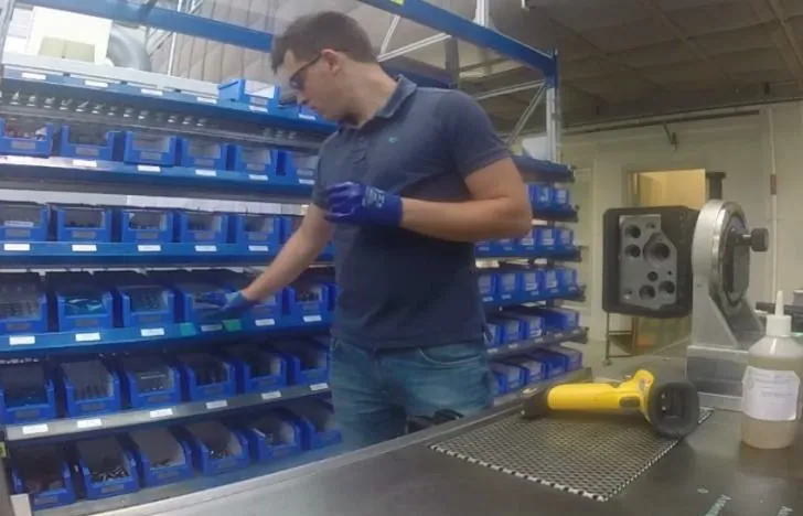 Operator picking components from a wall of blue picking bins next to a workbench with a barcode scanner